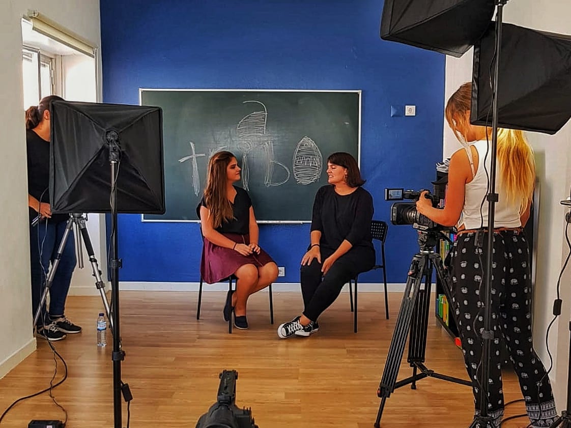 Entrevista numa sala de parede azul e quadro de ardósia, com duas softboxes e a câmara em tripé.